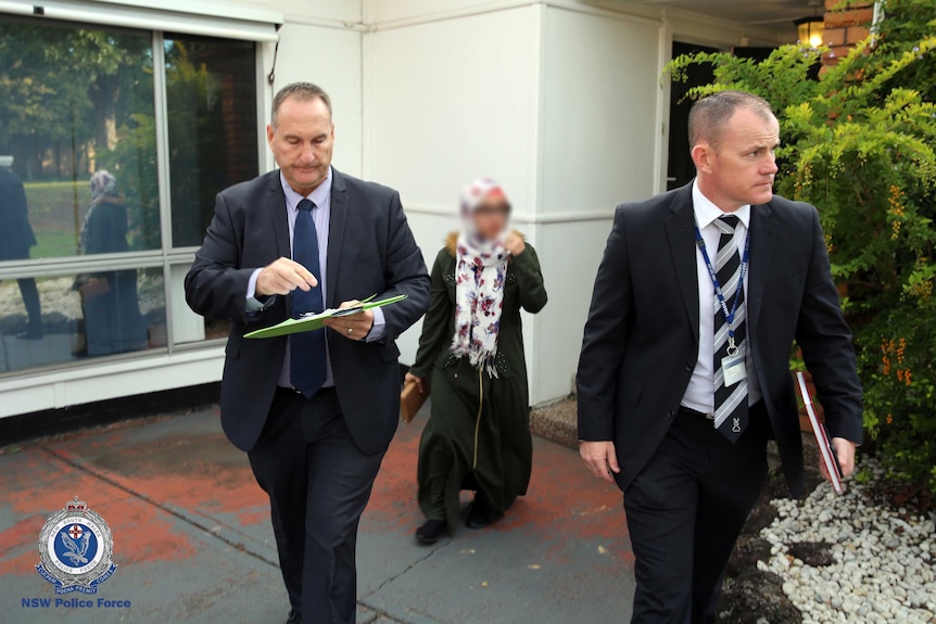 Two detectives walk out of a house as a lady with a blurred face follows