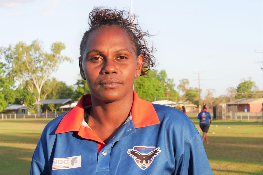 A woman wearing a team polo on a football oval looks at the camera 