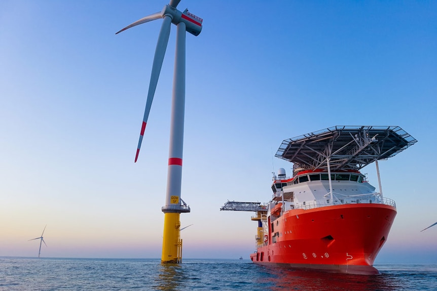 A red and white ship in the water next to a wind turbine. 