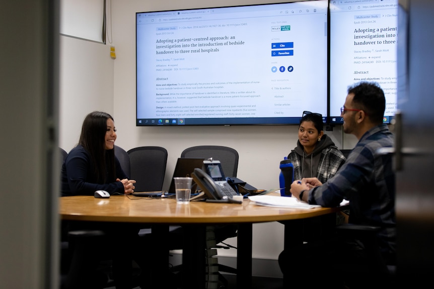 Three students sat in a conference room with a large screen behind them.
