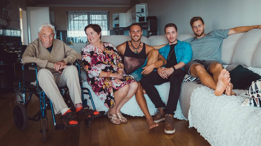 An old man in a wheelchair next to a couch with a woman and three young men sitting on it.