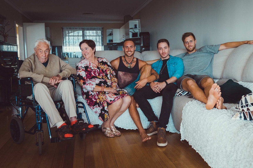 An old man in a wheelchair next to a couch with a woman and three young men sitting on it.