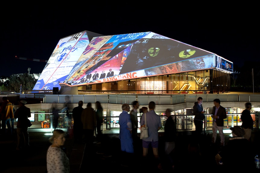 The exterior of a buildings triangular wall is lit up with a montage of images from past shows