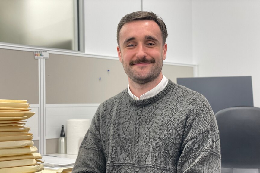 A man with short hair and mustache sits in an office wearing a grey jumper.