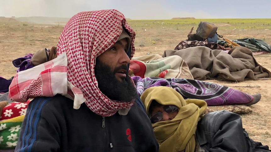 A man in a head scarf sits on the ground holding gauze while another man lies beside him.