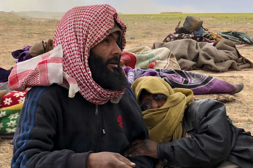 A man in a head scarf sits on the ground holding gauze while another man lies beside him.