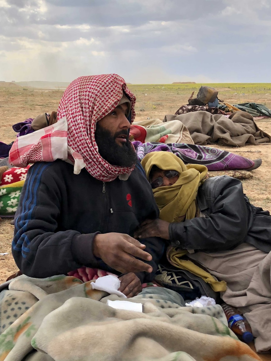 A man in a head scarf sits on the ground holding gauze while another man lies beside him.