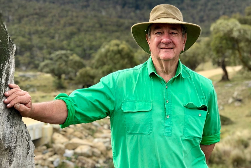 A man in jeans, a green shirt and a broad-brimmed hat leans up against an old tree.