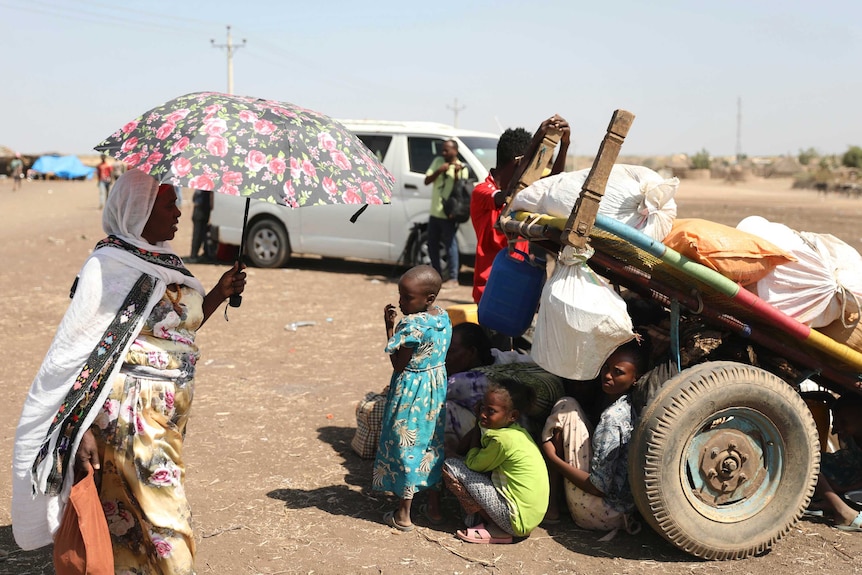 A woman holds an umbrella and people sit under a trailer to shield from the sun