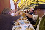 A group of people smile and clink their beers together over meals at Oktoberfest.