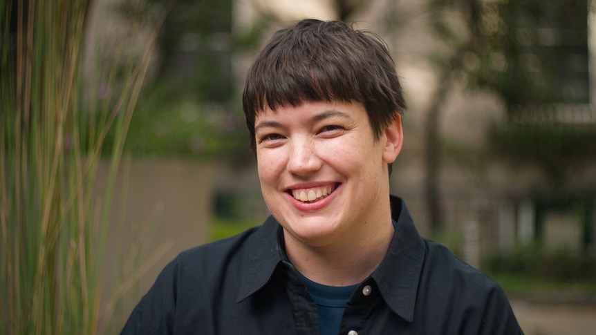 A smiling woman with a short haircut and straight fringe smiles outdoors.