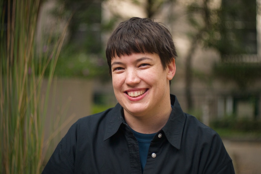 A smiling woman with a short haircut and straight fringe smiles outdoors.