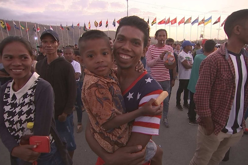 A smiling man holds a small boy in a crowd of people surrounded by flag poles