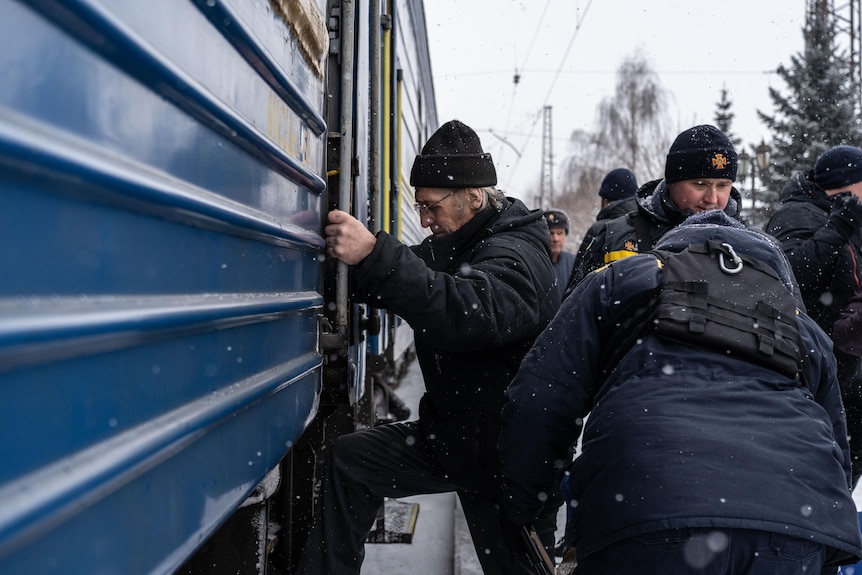 An old man in a big jacket and beanie clutches the outer sides of a train compartment as he steps in. Snow falls around him