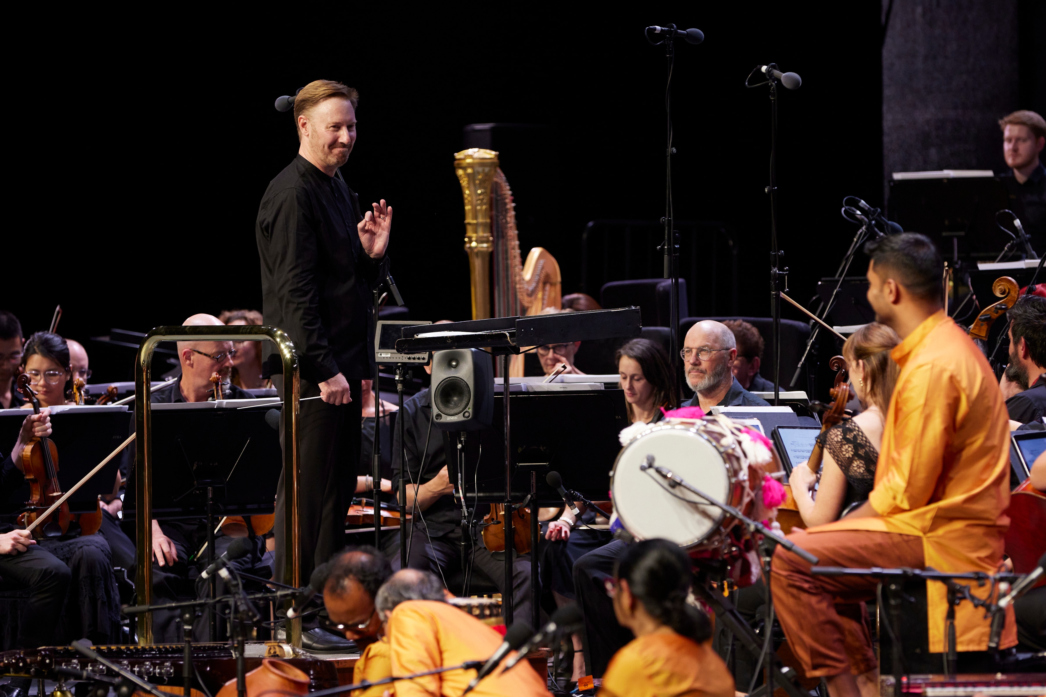 Benjamin Northey stands on a conductor's podium facing toward and Indian music ensemble smiling and giving them an ok sign.