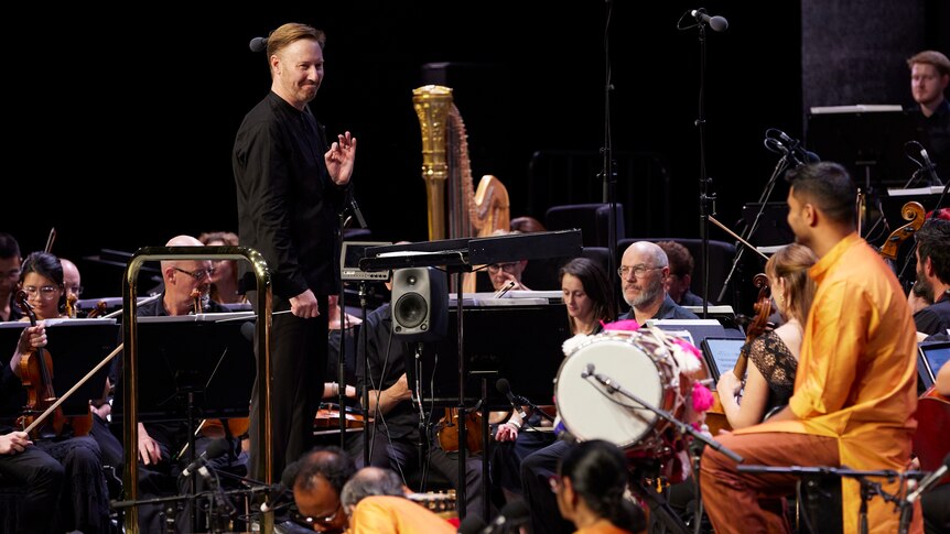 Benjamin Northey stands on a conductor's podium facing toward and Indian music ensemble smiling and giving them an ok sign.