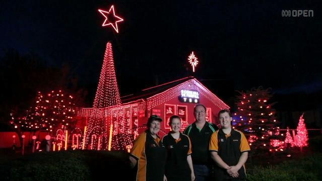 Four people stand in front of a house full of Christmas lights at night