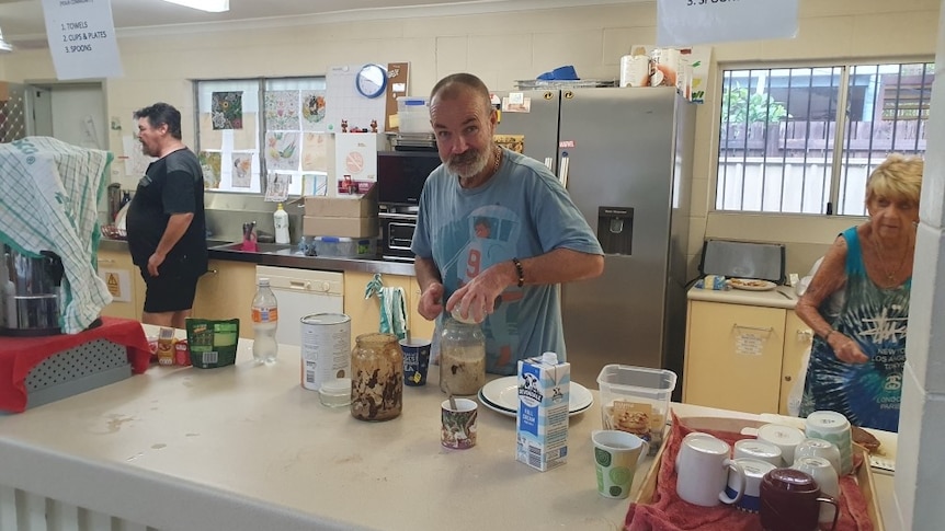 A man makes a cup of coffee while others stand around in kitchen of community drop-in centre.