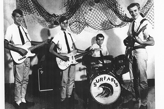 A group of four young men in shirts and ties, three holding guitars and one at a drum kit with the word 'Surfaris' on the front