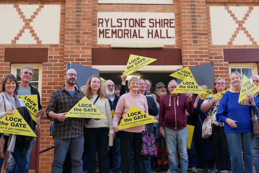 People standing in front of a hall with the sign "Rylstone Shire Memorial Hall" holding yellow signs that say "lock the door"