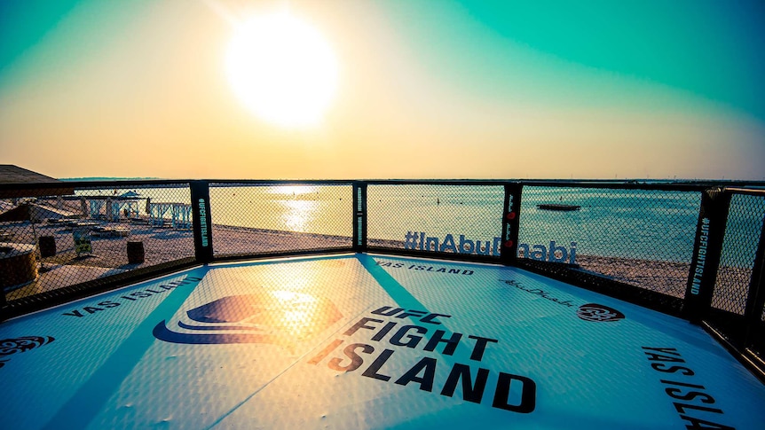 A UFC octagon, with "Fight Island" written on it, on the beach at Yas Island. The sun is setting.