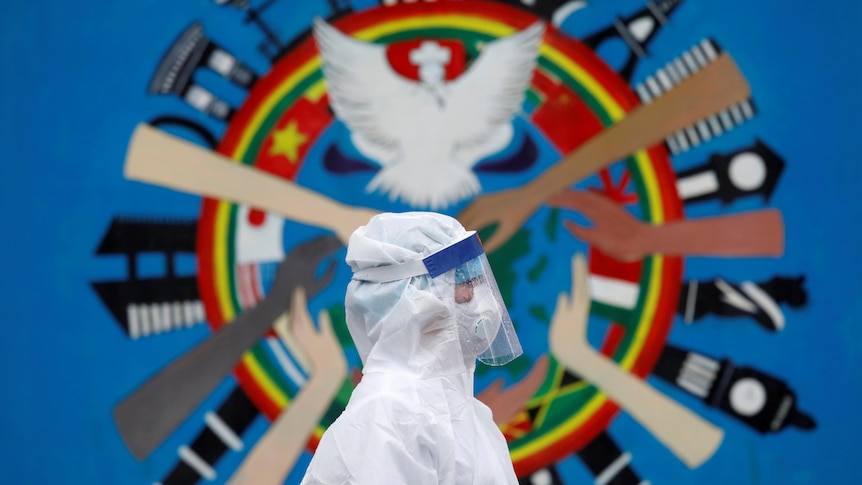 A person in white health PPE suit in front of a circular mural with painted hands and a white dove