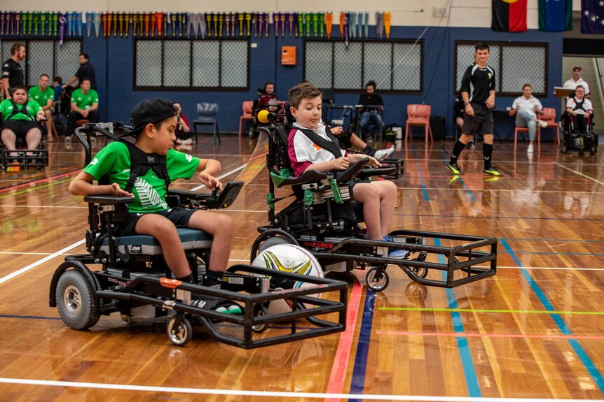 Two boys in electric wheelchairs chase a ball that is rolling between them