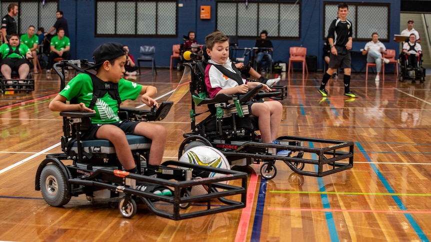 Two boys in electric wheelchairs chase a ball that is rolling between them