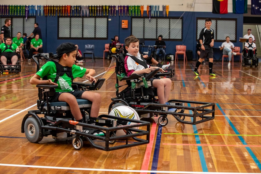 Two boys in electric wheelchairs chase a ball that is rolling between them