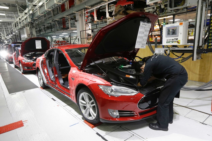 A Tesla employee bends down to work on the motor of a Model S at the front of a line of cars.