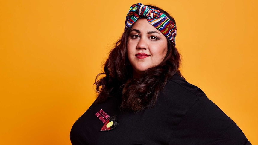 Comedian Steph Tisdell standing in front of a mustard coloured background.