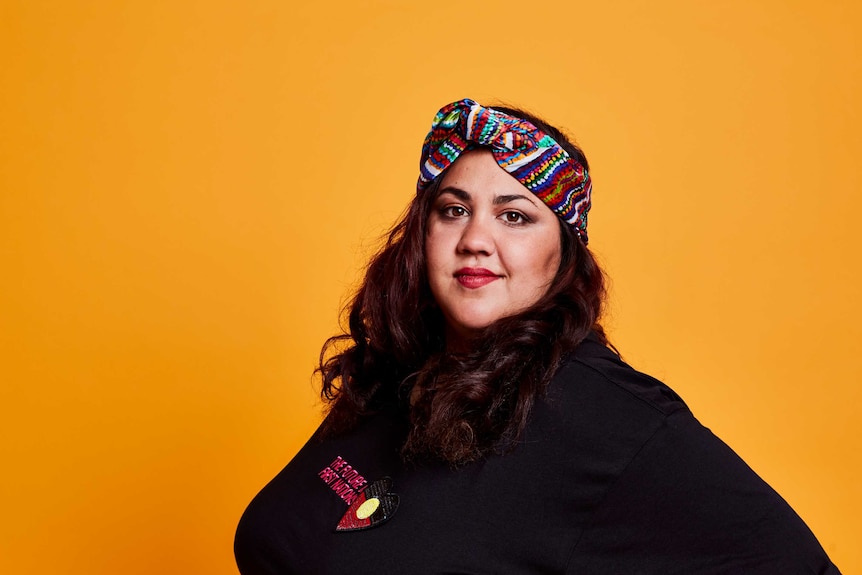 Comedian Steph Tisdell standing in front of a mustard coloured background.