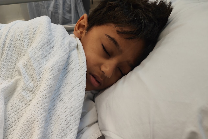 Young boy with dark skin and hair lies in a white hospital bed, tucked in by a sheet.
