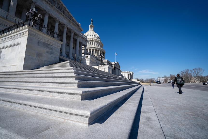 The steps that lead to the US Capitol building are in focus, a man in a suit walks past with back to camera
