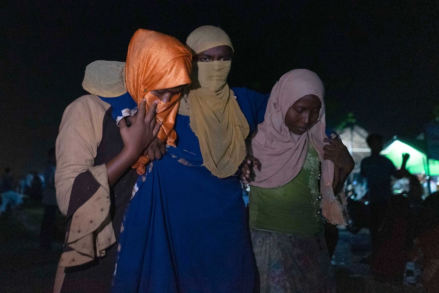 Two women assist another woman to walk at night time. All three wear head coverings.