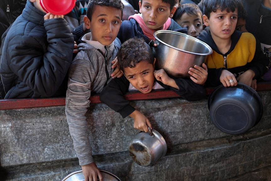 A young boy yells as other children with empty bowls crowd around a barrier where food is being prepared