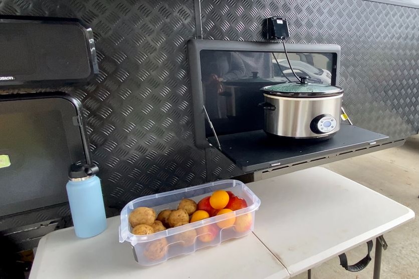 A plastic container with vegetables sits on a white table beside a caravan with a cooking pot on a ledge.