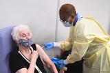 An older woman wearing a floral mask sits in a chair as a healthcare worker in PPE gives her a needle