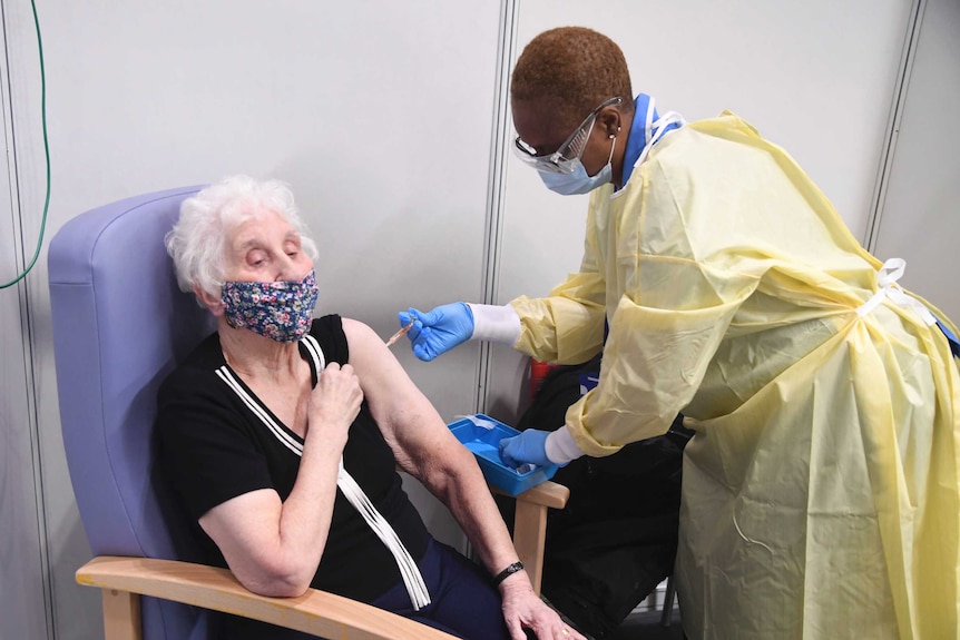 An older woman wearing a floral mask sits in a chair as a healthcare worker in PPE gives her a needle