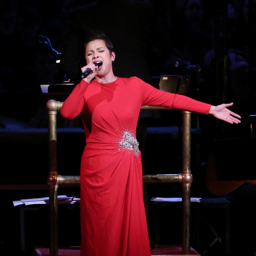 Lea Salonga in a red dress stands on stage in front of an orchestra and sings with eyes closed into a microphone.