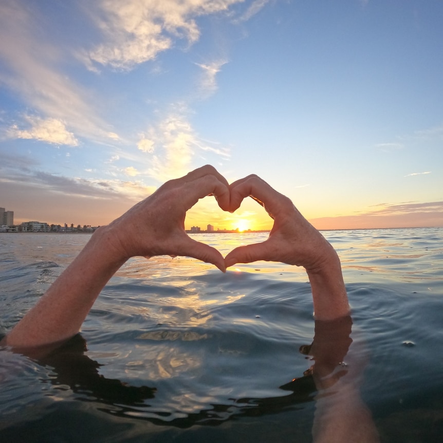 A person holding their hands out of the water and making a heart with the sun rising.