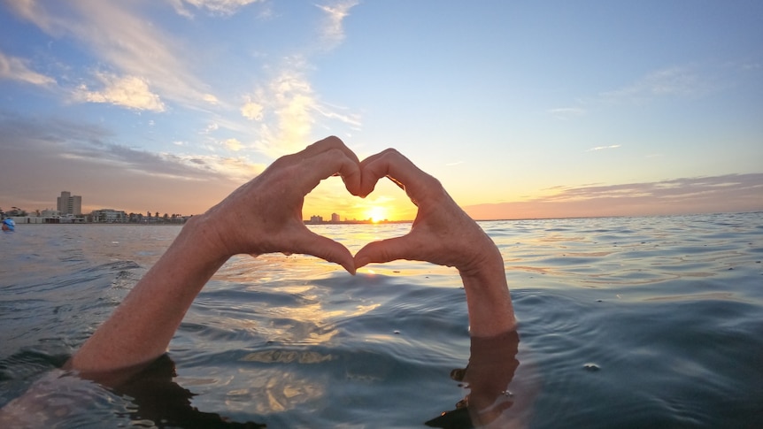 A person holding their hands out of the water and making a heart with the sun rising.