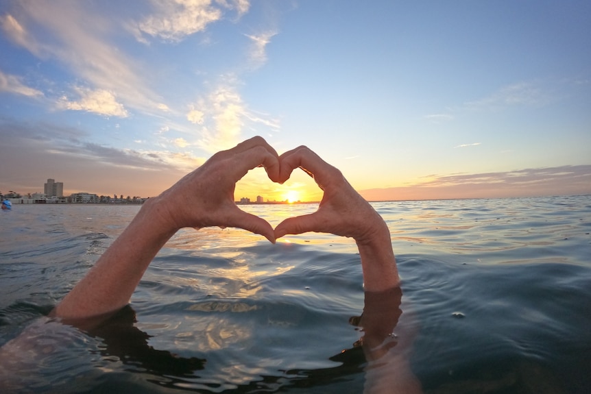 A swimmer's hands form a love heart through which the sun shines at rises over a city.
