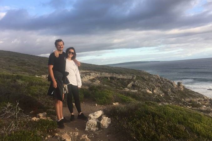 A young man and young woman stand arm in arm near the coast.