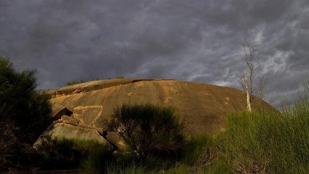 Large rock formation under stormy sky, with bushes at its base