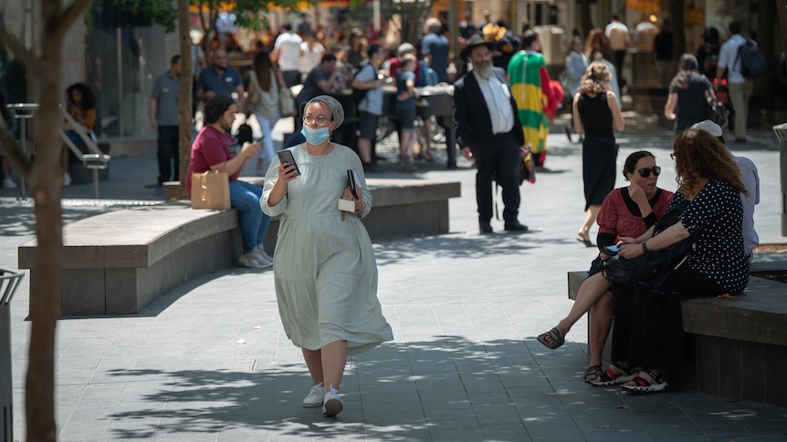 A woman wearing a knee-length dress and scarf covering her hair walks down the street looking at her phone