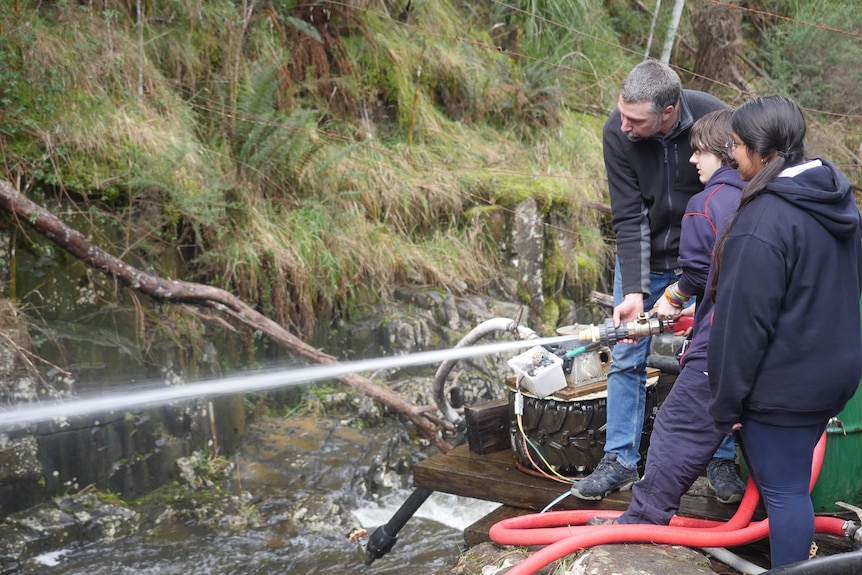 A man and two school students hold onto and use a high pressured hose in a rocky creek