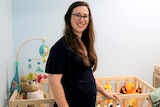 A pregnant lady stands in front of a cot smiling