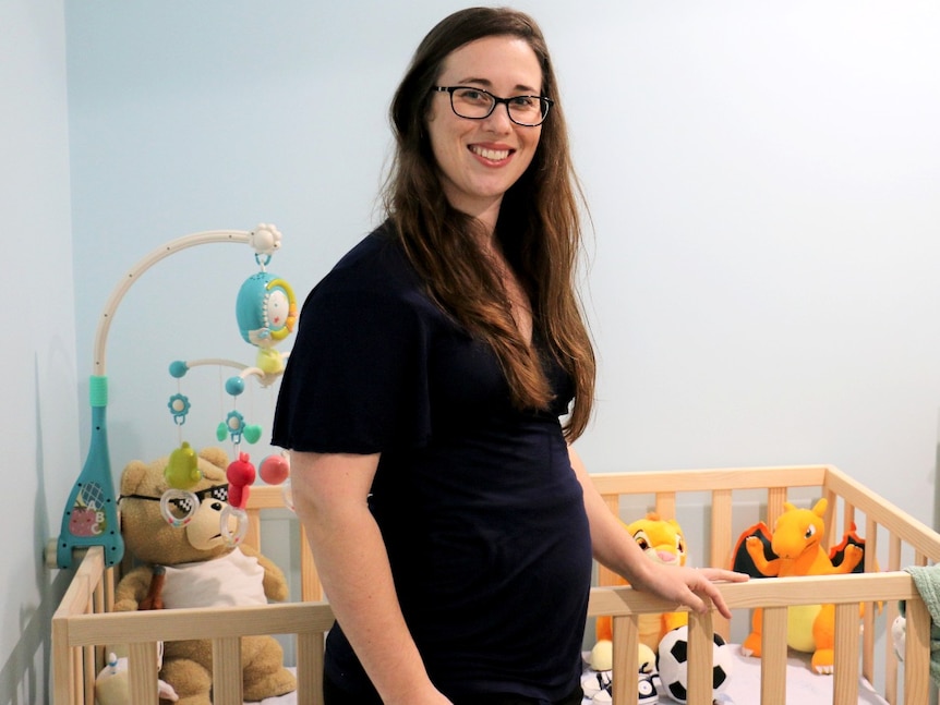 A pregnant lady stands in front of a cot smiling