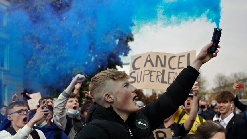 A young male in a crowd holds up a flare eminating blue smoke. A sign behind him reads "CANCEL SUPERLEAGUE"